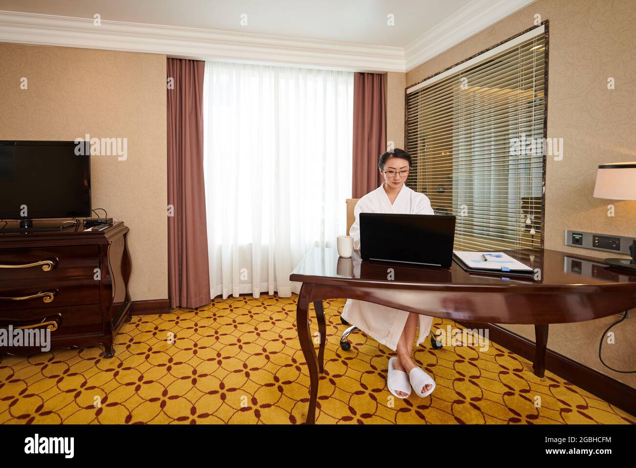 Konzentrierte junge asiatische Geschäftsfrau im Hotelbademantel, die während der Arbeit im Urlaub auf dem Laptop tippt Stockfoto