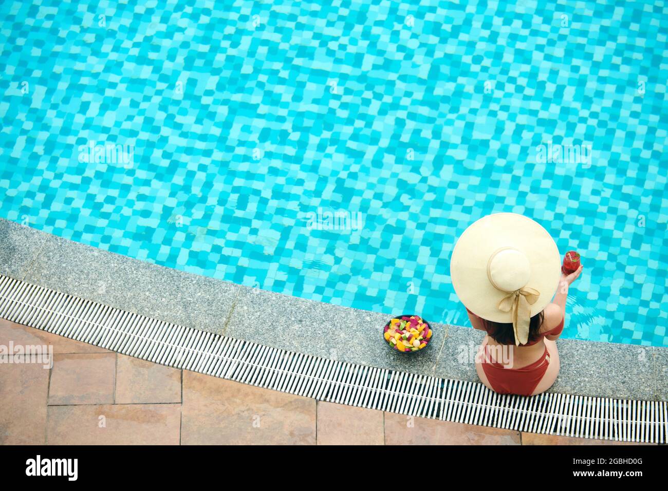 Direkt über der Ansicht einer nicht erkennbaren Frau in Strohhut und roter Bademode, die Cocktail trinkt und in der Nähe des Pools sitzt Stockfoto