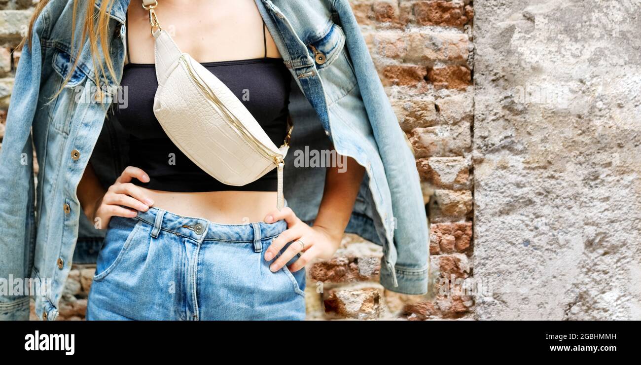 Junge schöne 3/4-Frau, die neben der Mauer aus Ziegelsteinen steht, Jeans-Kleidung aus Denim, Gürteltasche in der Schulter, Sommer-Modetrend, Stockfoto