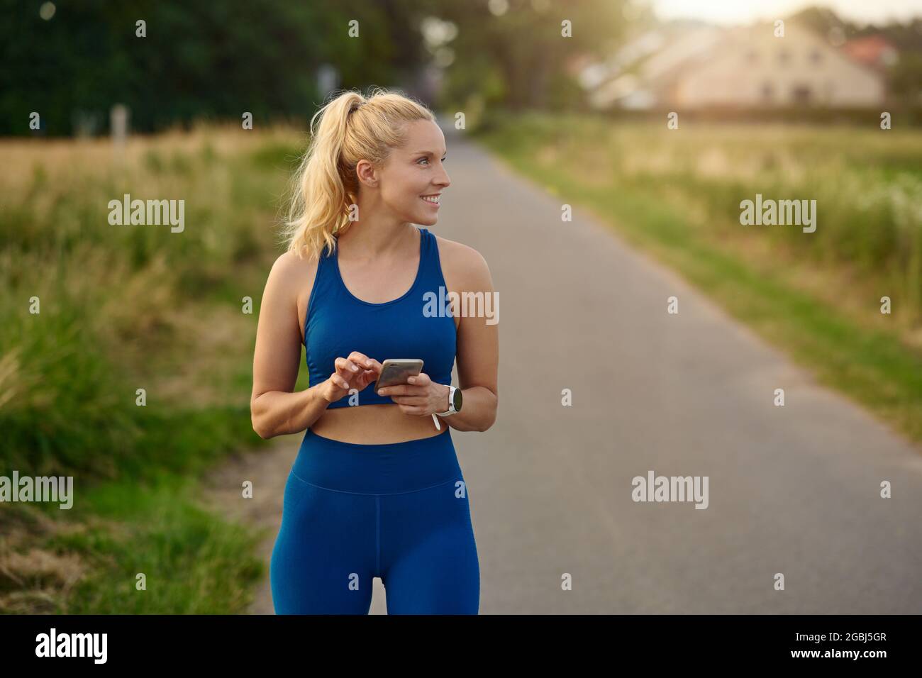 Fit athletische Frau pausend, um ihr Handy zu konsultieren, während Sie joggen auf einem ländlichen Wanderweg Blick zur Seite mit einem glücklichen Lächeln der Zufriedenheit und Stockfoto