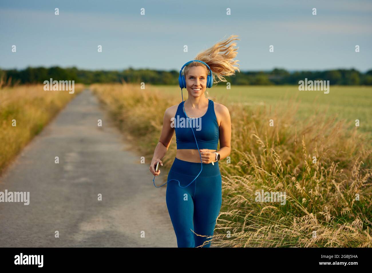 Fit temperamentvolle junge Frau joggt auf einem Fußweg in offenen Feldern und nähert sich der Kamera mit einem glücklichen Lächeln und Pferdeschwanz, der in einer Gesundheit nach hinten fliegt Stockfoto