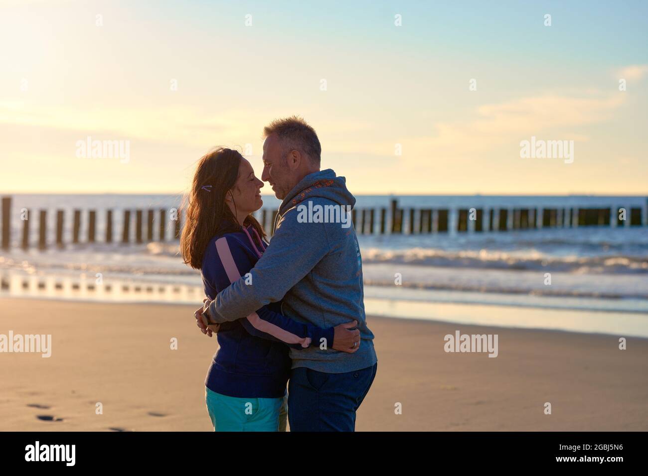 Ein romantisches Paar mittleren Alters, das sich bei Sonnenuntergang an einem tropischen Strand umarmt und sich mit seitlichem Copyspace liebevoll in die Augen schaut Stockfoto