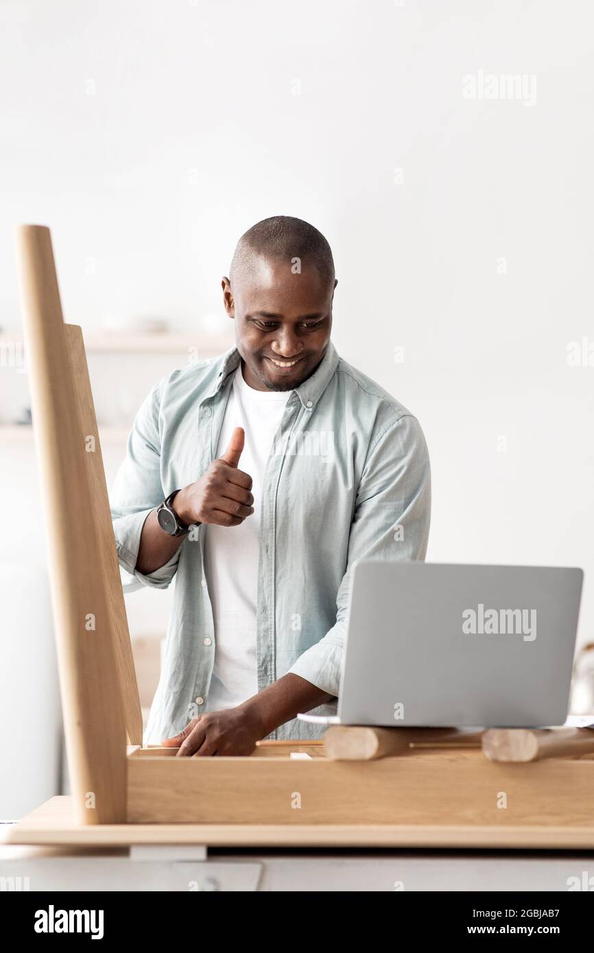 Blog über Möbelmontage. Positiver afroamerikanischer Mann, der den Daumen auf den Laptop zeigt, Platz kopieren, ausschneiden Stockfoto