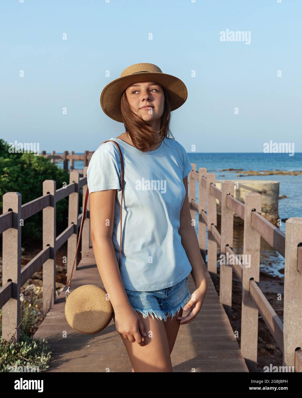 Das Teenager-Mädchen steht bei Sonnenuntergang auf einem Holzsteg am Meer und trägt ein hellblaues T-Shirt, einen Strohhut und eine Geldbörse. Reisekonzept für den Sommer Stockfoto