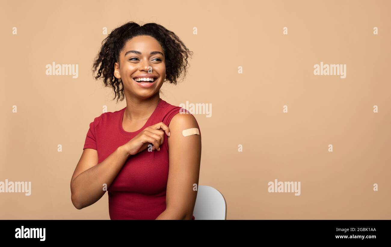 Fröhliche geimpfte schwarze Frau, die nach der Injektion des Coronavirus-Impfstoffs auf beigem Hintergrund einen Arm mit Verband zeigt Stockfoto