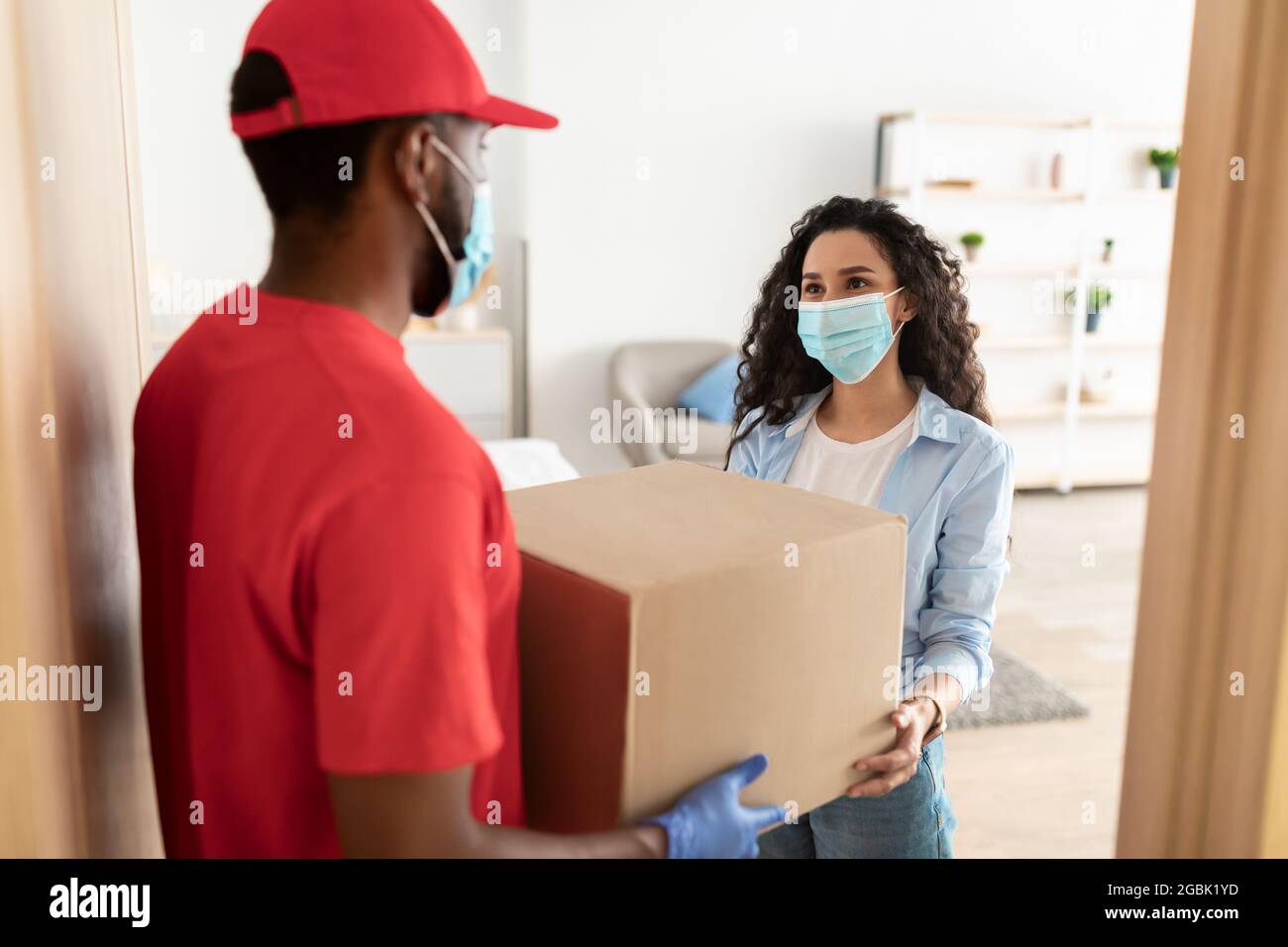 Schwarzer Deliveryman in Einwegmaske geben Box an Frau Stockfoto