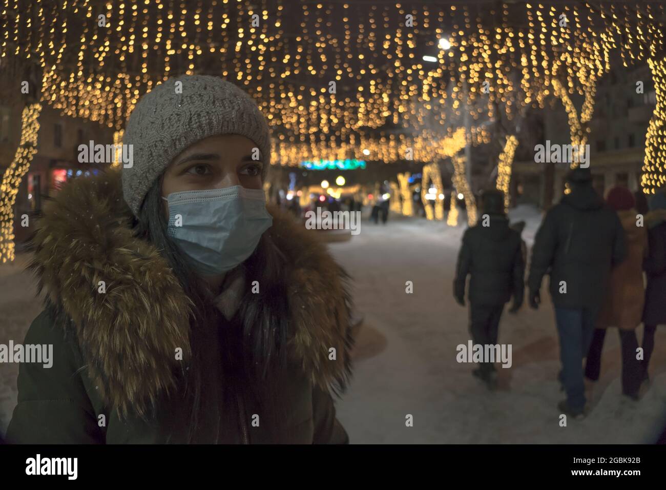 Ein Mädchen in einer medizinischen Maske auf der Neujahrsstraße. Eine Frau in einer Gesichtsmaske steht auf einer Straße mit Geschäften, die für Weihnachten und Neujahr dekoriert sind. Urlaubsdauer Stockfoto