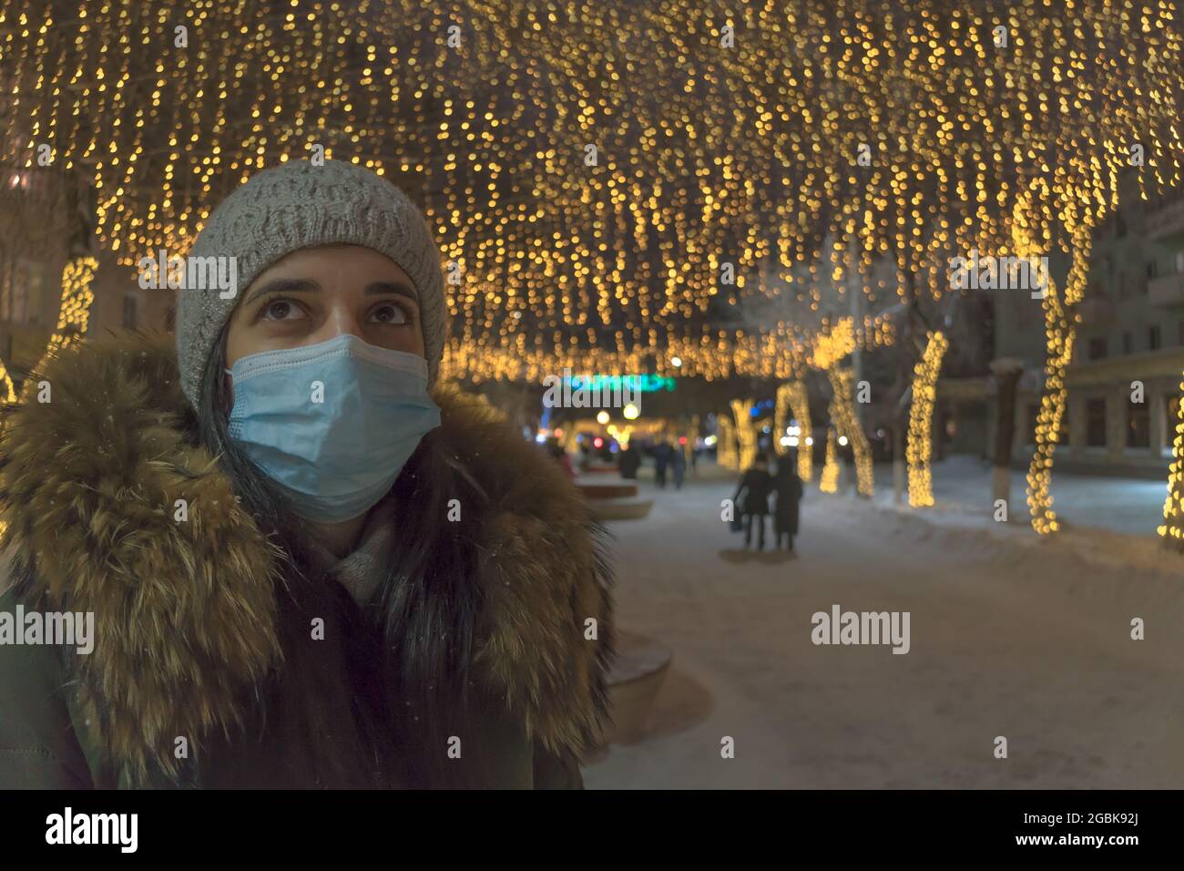 Ein Mädchen in einer medizinischen Maske auf der Neujahrsstraße. Eine Frau in einer Gesichtsmaske steht auf einer Straße mit Geschäften, die für Weihnachten und Neujahr dekoriert sind. Urlaubsdauer Stockfoto