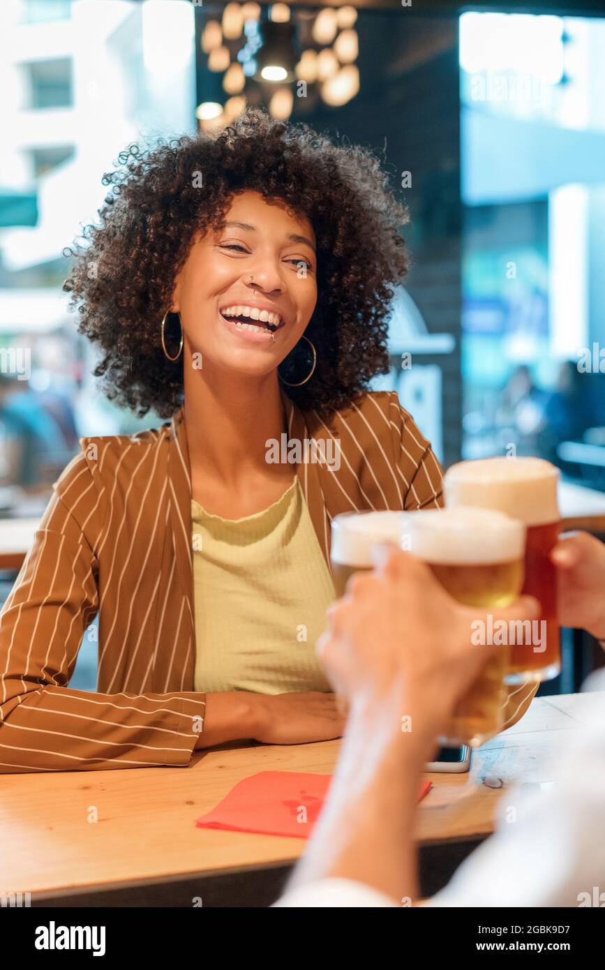 Fröhliche, lebhaft lachende, junge Schwarze Frau, die mit einem kalten Bier toast, während sie mit einem Freund in einem Pub oder Restaurant in einer Stadtstraße feiert Stockfoto