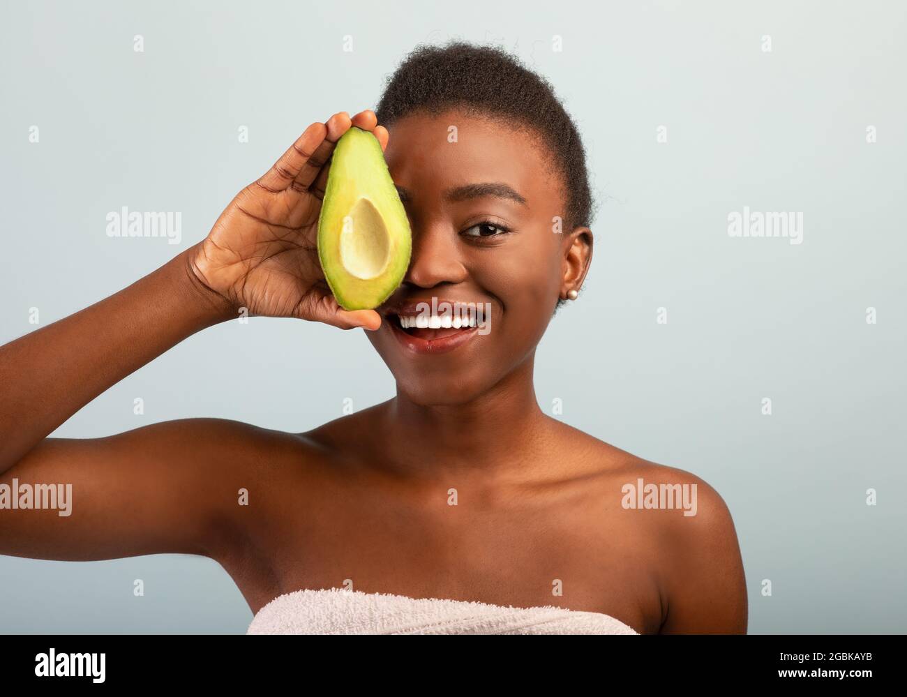 Glückliche afroamerikanische junge Frau, die frische Avocadohälften in der Hand hält und das Auge bedeckt, lächelt vor der Kamera auf grauem Hintergrund Stockfoto