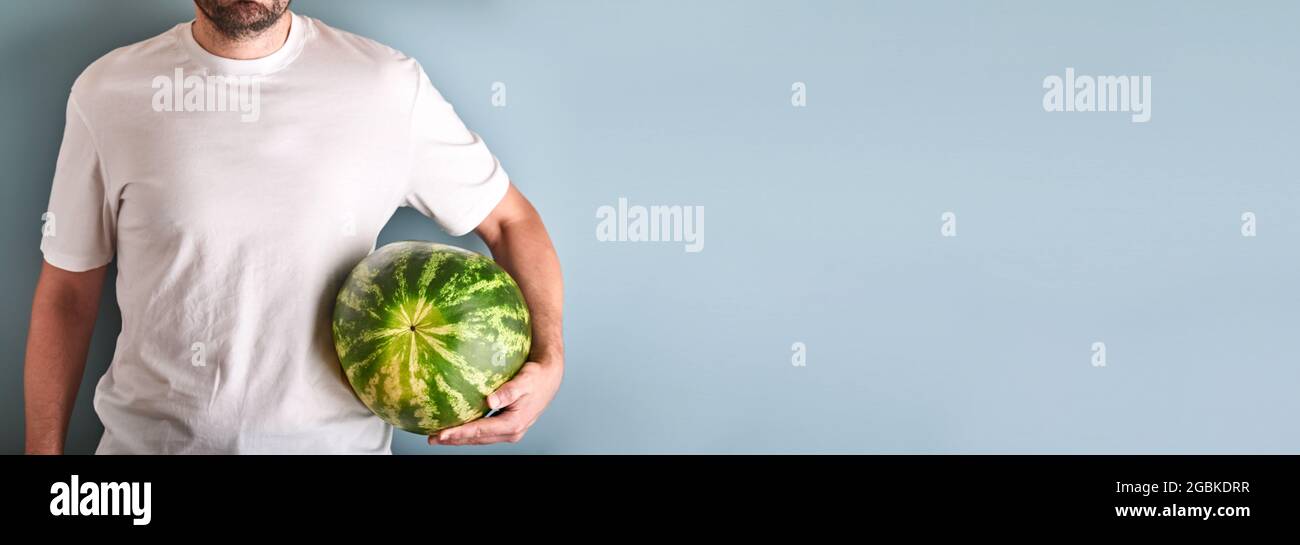 Junger Mann hält ganze Wassermelone in den Händen in weißem T-Shirt auf blauem Hintergrund. Design-Modell. Speicherplatz kopieren. Stockfoto