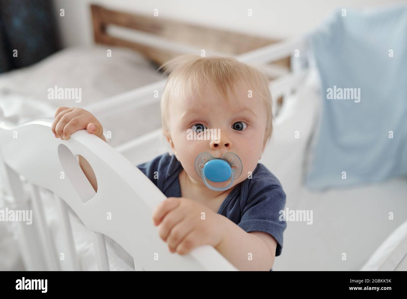 Porträt eines neugierigen blonden Babys mit Schnullerfigur, die die Gitterseite im Schlafzimmer mit einer Gitterrostwand hält Stockfoto