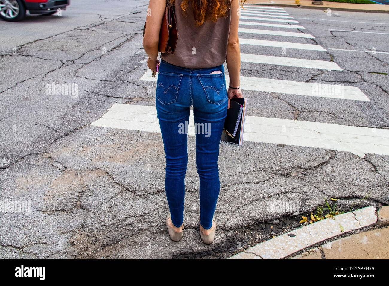 Zurück in die Schule - nicht identifizierbare Rückseite des Mädchens in Jeans mit langen roten Haaren, die auf dem Fußgängerüberweg mit Notizbüchern und einer Ledertasche und Schlüsseln stehen Stockfoto