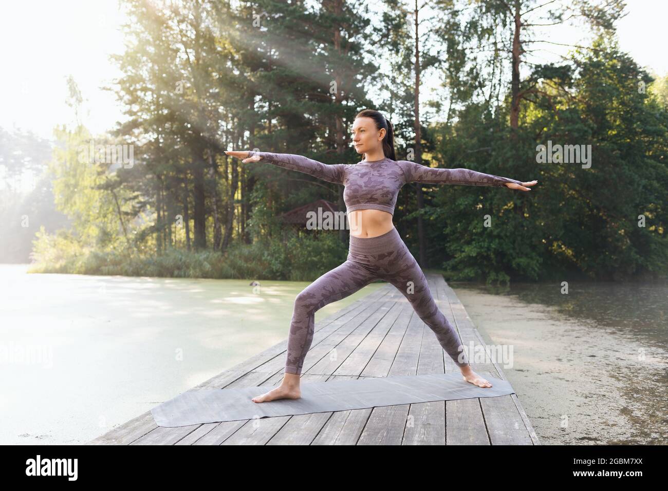 Eine Frau, die am Morgen auf einer Holzbrücke im Park Yoga praktiziert, führt virabhadrasana, eine Kriegerpose, durch Stockfoto