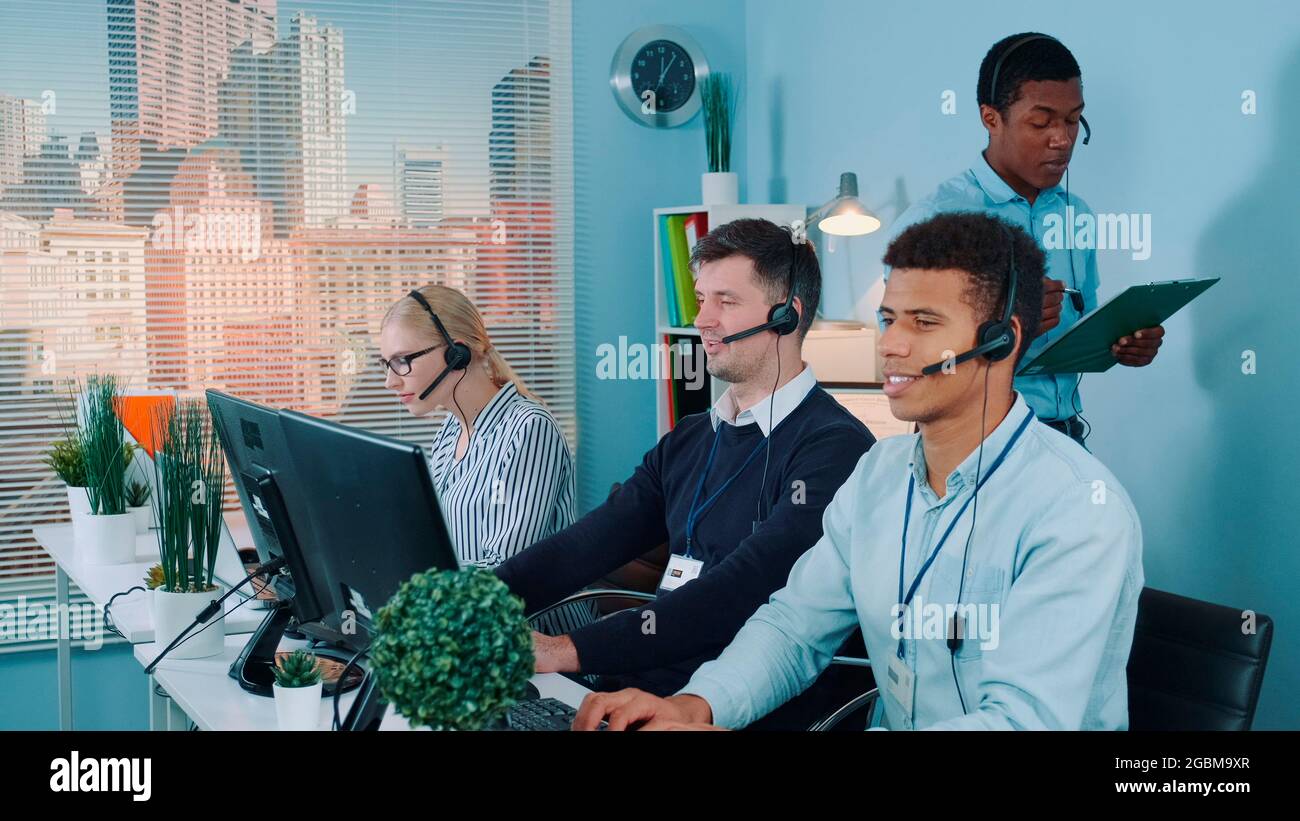 Ein multiethnischer Kundendienstmitarbeiter, der seinen Kollegen einen Witz erzählt, während er die Kunden anruft. Sie lachen und arbeiten weiter. Stockfoto