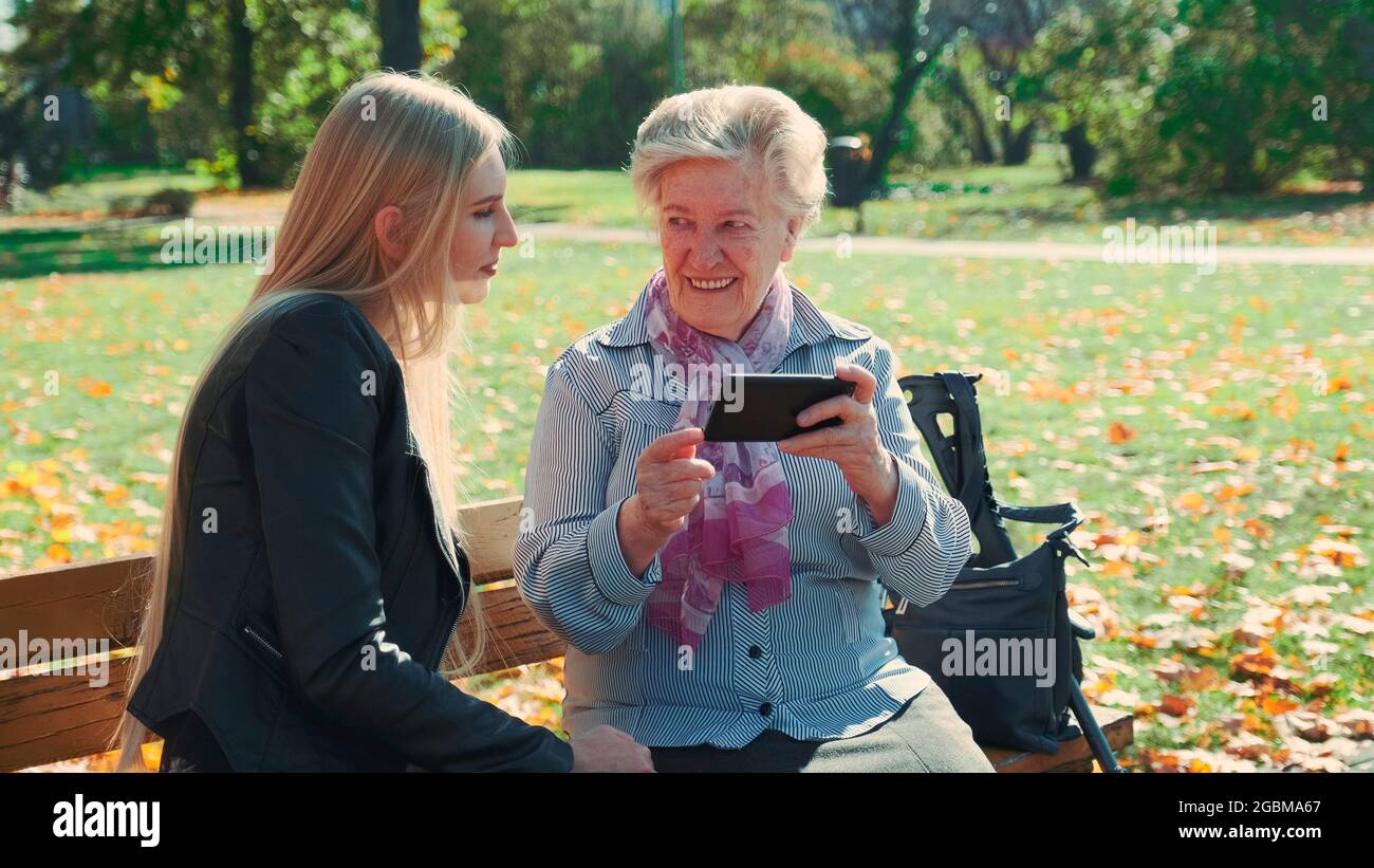 Ältere Großmutter, die ihre Enkelin auf dem Smartphone zeigt. Sie sitzen auf der Bank und reden im Park. Stockfoto