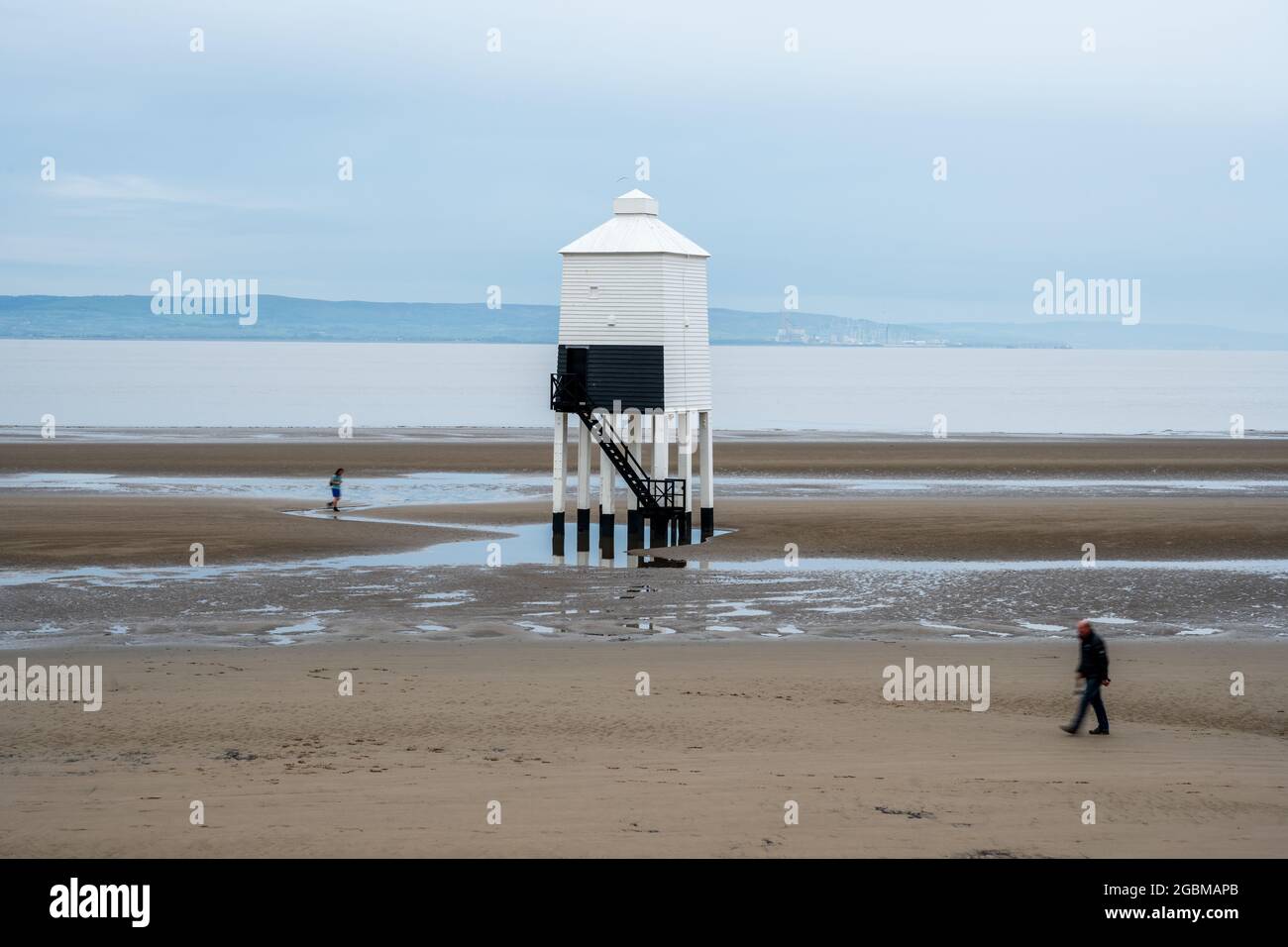 Ein Jogger und Spaziergänger passieren den niedrigen Leuchtturm am Burnham-on-Sea Beach in Somerset, mit Bridgwater Bay, Hinkley Point und dem Quantock Hills Behi Stockfoto