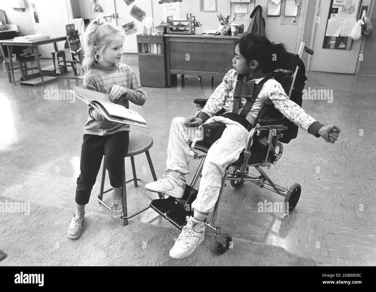 Austin Texas USA, um 1989: Das Mädchen der vierten Klasse hält ein Buch und liest im öffentlichen Schulzimmer einer rollstuhlgebundenen Klassenkameradin mit zerebraler Lähmung vor. KEINE Releases. ©Bob Daemmrich Stockfoto