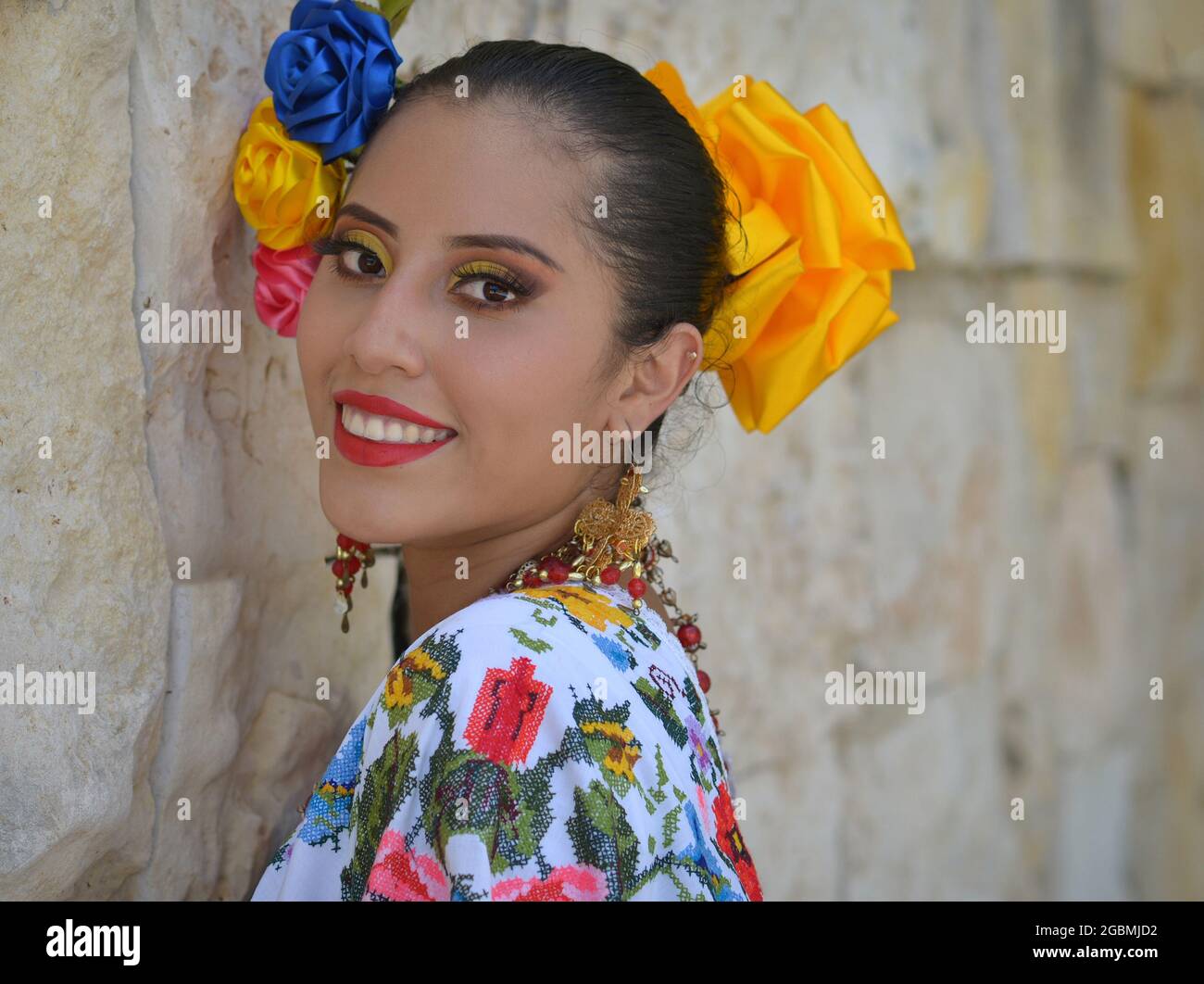 Die junge schöne mexikanische Yucatecan-Volkstänzerin trägt ein traditionelles folkloristisches Kleid mit bunten Blumen im Haar und lächelt für die Kamera. Stockfoto