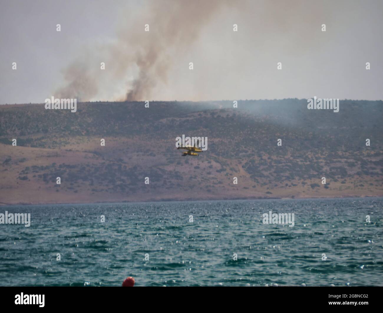 Blick auf einen gelben Hubschrauber, der auf das Meer fällt und auf einen unbefestigten Berg fällt und im Hintergrund Rauch aufgeht Stockfoto