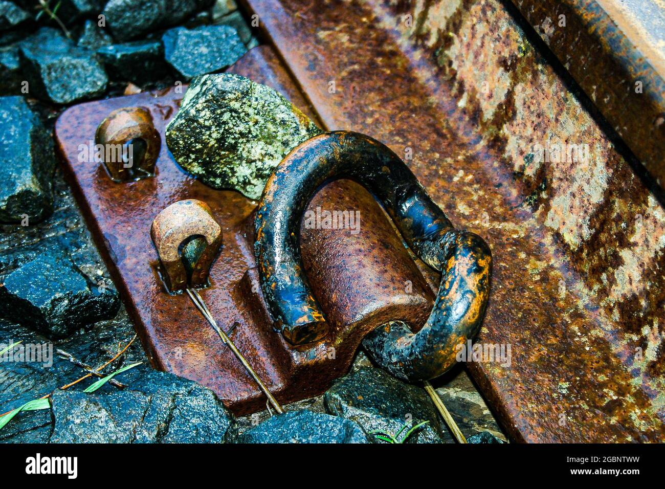 Nahaufnahme einer verwitterten, rostigen Klammer, die die Eisenbahnstrecke zum Schläfer hält, der auf dem Ballast sitzt Stockfoto