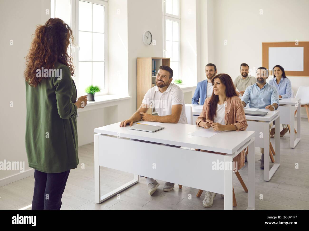 Business Coach im Gespräch mit glücklichen erwachsenen Studenten, die an Schreibtischen im Klassenzimmer sitzen Stockfoto