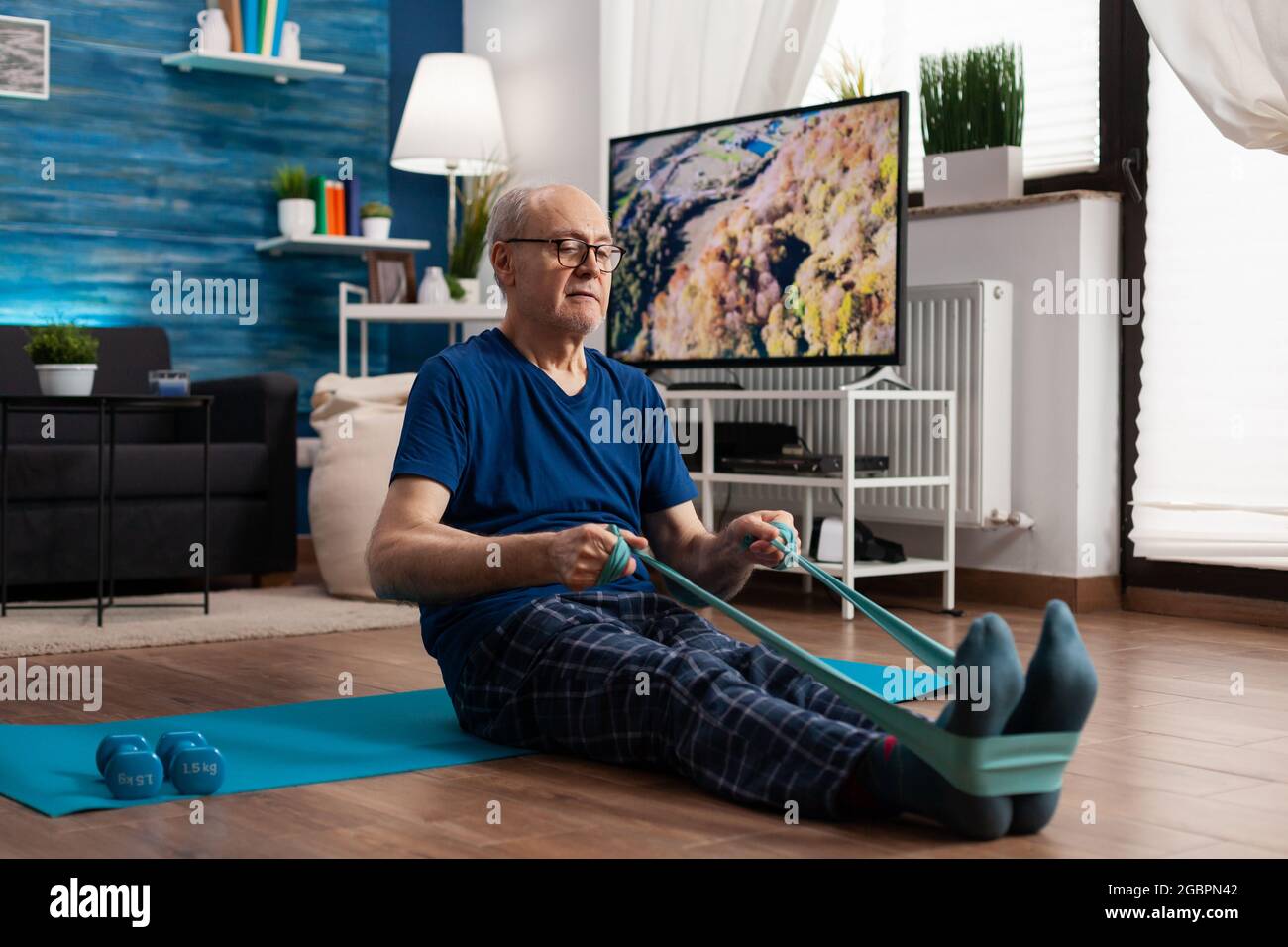 Seniorchef im Ruhestand sitzt auf einer Yogamatte und dehnt die Beine mit einem elastischen Band aus, um die Körperflexibilität zu trainieren. Rentner in Sportbekleidung abnehmen Gewicht beim Muskeltraining im Wohnzimmer Stockfoto