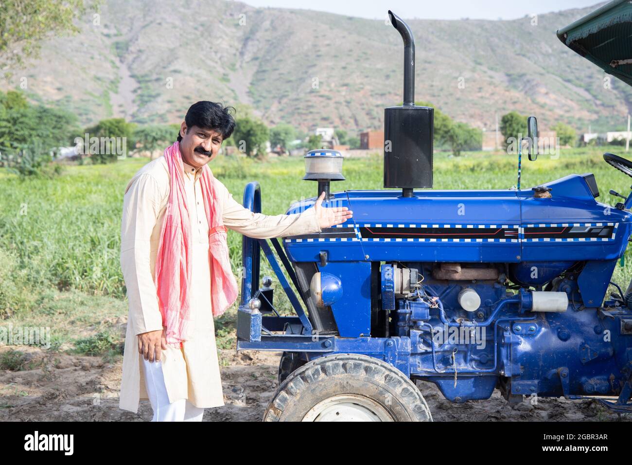 Junger, glücklicher indischer Bauer, der seinen blauen Traktor auf dem Agrarfeld zeigt. Mann in traditioneller Kurta mit lächelndem Schnurrbart und Blick auf den Kamerafelder Stockfoto