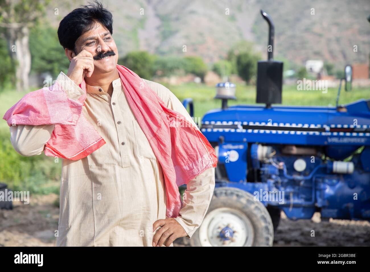 Junger, glücklicher indischer Bauer berührt seinen Schnurrbart, der auf dem Landwirtschaftsfeld steht, mit einem blauen Traktor hinter ihm. Der Mann, der traditionelle Kurta trägt, fühlt sich stolz. Stockfoto