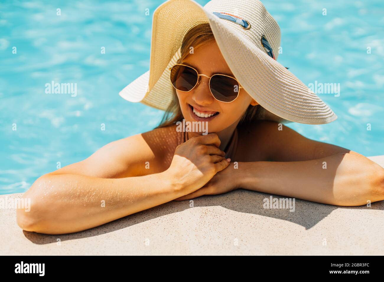 Schöne junge gebräunte Frau in Sonnenbrille und Sommerhut, im Außenpool, Frau in einem Badeanzug Sonnenbaden im Pool, glückliche Frau in Th entspannen Stockfoto