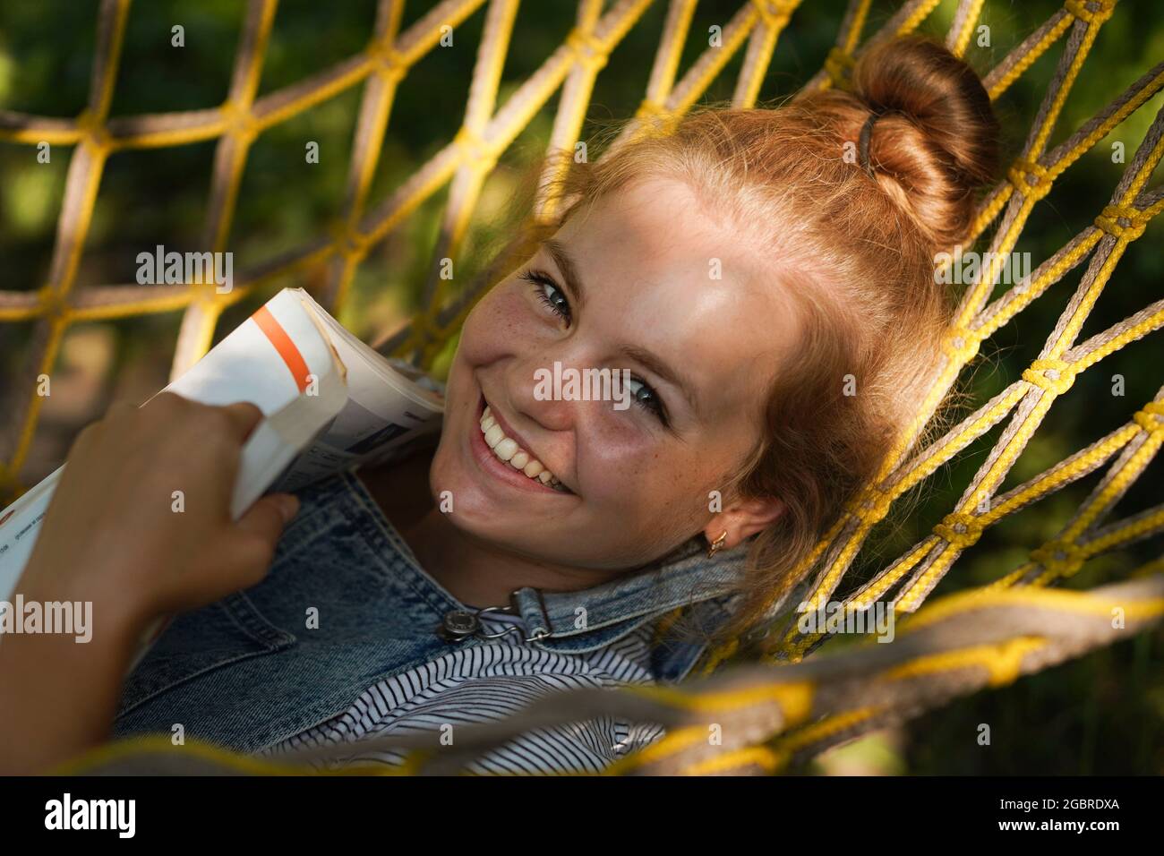 Junge blonde Studentin, die lächelt und ein Buch liest, während sie auf EINER Hängematte im Garten liegt. Frau, die sich in der Hängematte entspannt. Zurück zur Schule, Universitätscampus Stockfoto