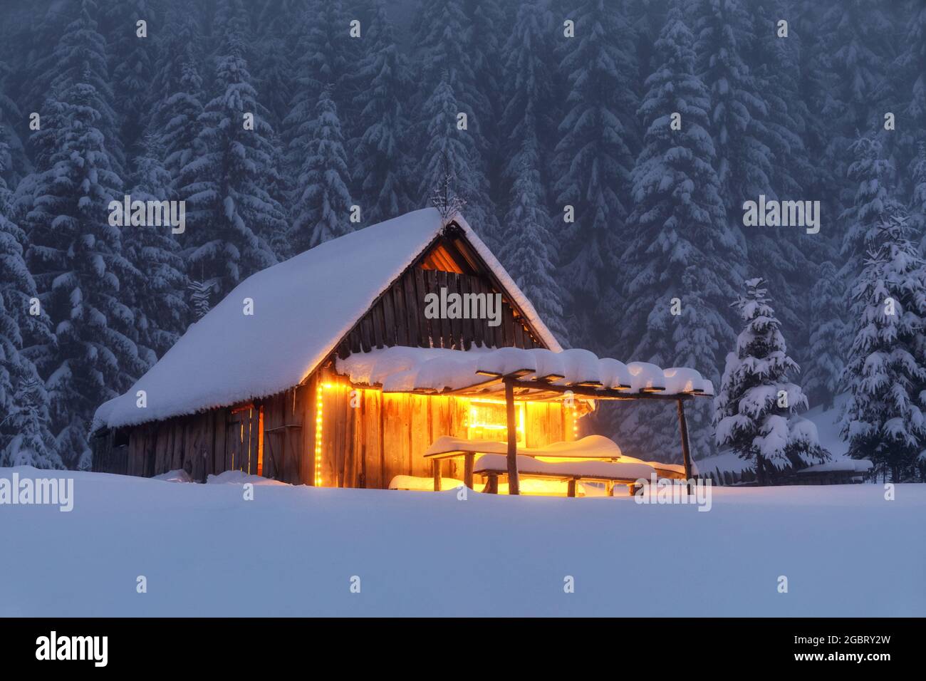 Holzhütte auf dem mit Schnee bedeckten Rasen. Abends leuchten die Lampen das Haus auf. Winterlandschaft. Mystische Nacht. Berge und Wälder. W Stockfoto