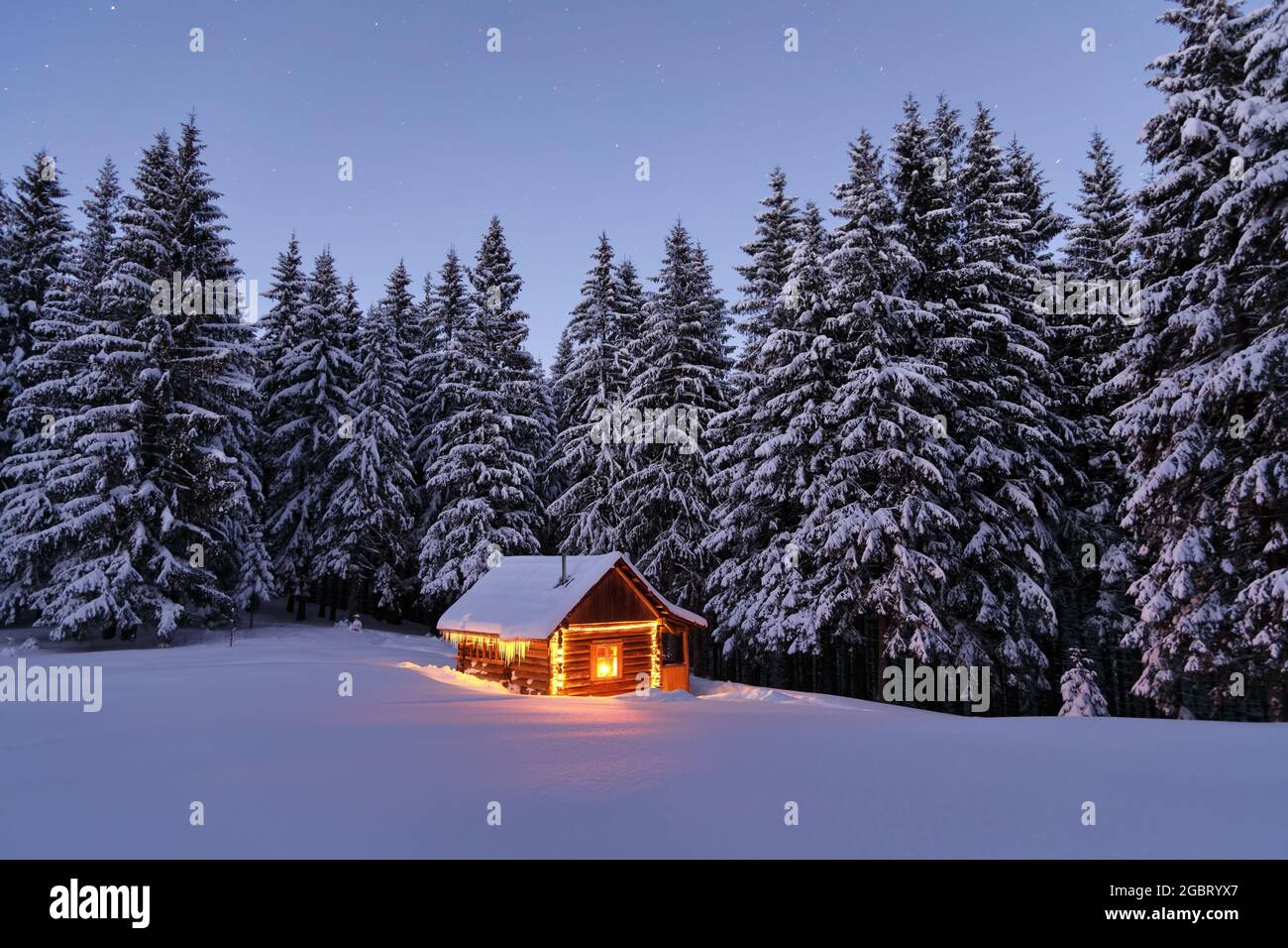 Holzhütte auf dem mit Schnee bedeckten Rasen. Abends leuchten die Lampen das Haus auf. Winterlandschaft. Mystische Nacht. Berge und Wälder. W Stockfoto