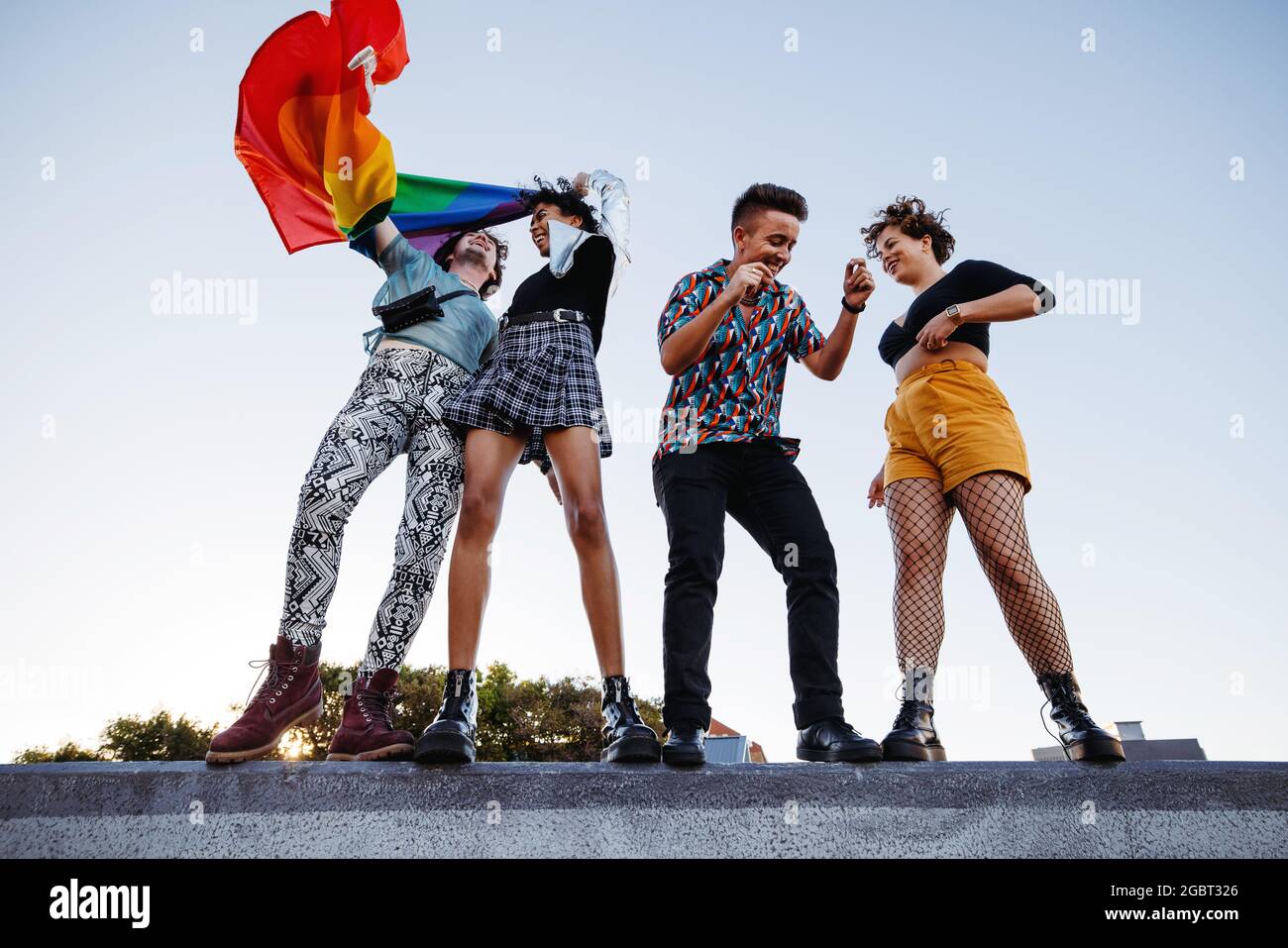 Glückliche Gruppe von queeren Individuen feiern Homosexuell Stolz zusammen. Vier Mitglieder der LGBTQ+ Community tanzen fröhlich und heben die Pride-Flagge. Stockfoto