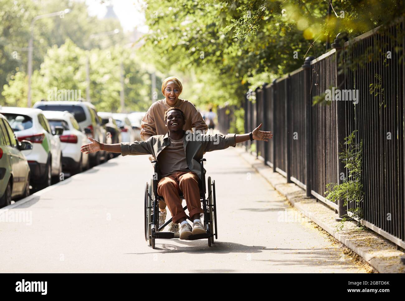 Glücklich aufgeregt jungen afroamerikanischen behinderten Mann im Rollstuhl von laufender Frau ausgestreckt Arme und das Gefühl der Freiheit gestoßen Stockfoto