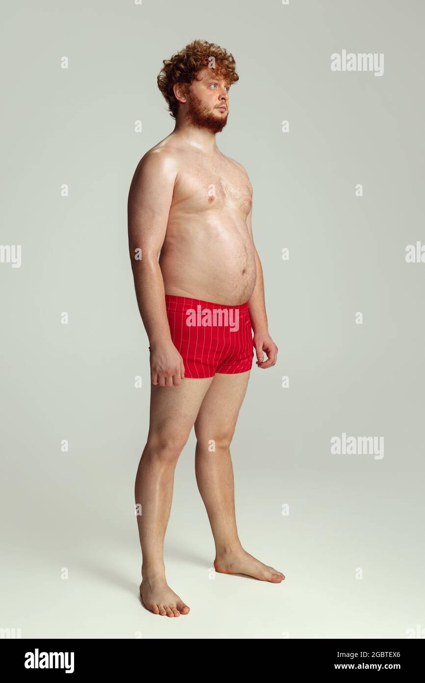 Niedlicher rothaariger Mann in roten Badeshorts, der isoliert auf grauem Studiohintergrund posiert. Konzept von Sport, Humor und Körper positiv. Stockfoto