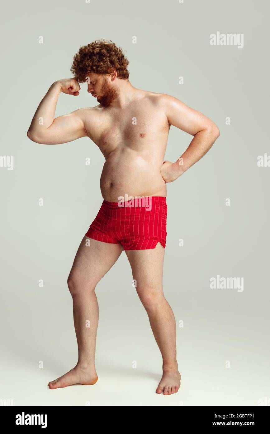 Niedlicher rothaariger Mann in roten Badeshorts, der isoliert auf grauem Studiohintergrund posiert. Konzept von Sport, Humor und Körper positiv. Stockfoto