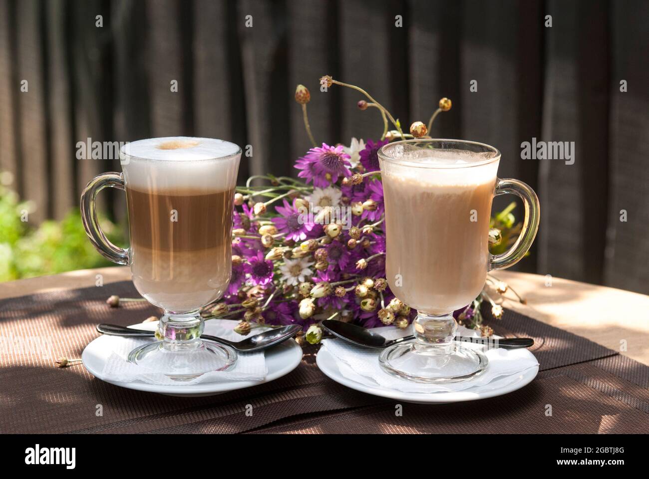 Ein Glas Latte und Kaffee glänzen auf dem Hintergrund der Blumen. Kaffeegetränke auf der Sommerterrasse des Restaurants Stockfoto