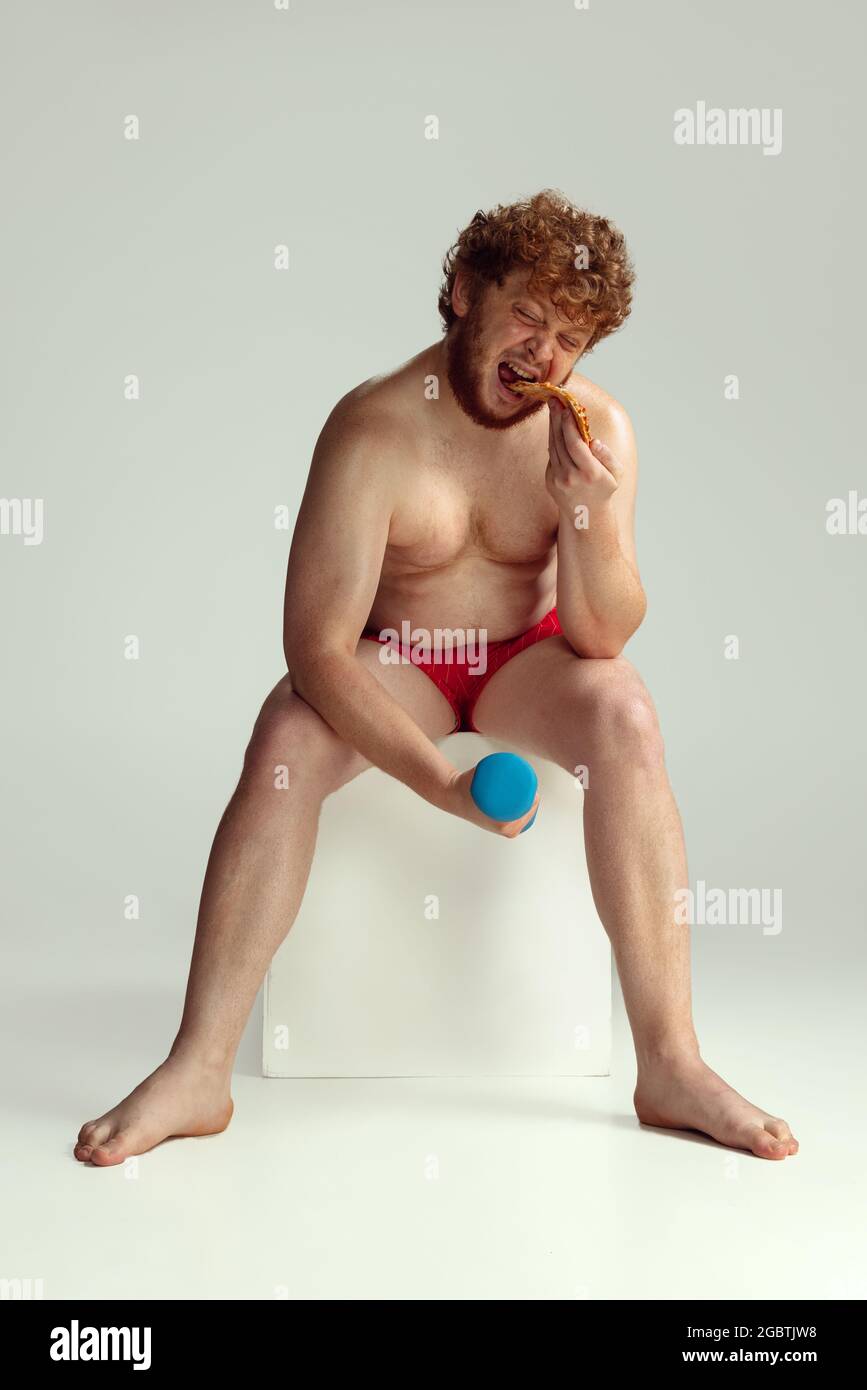 Niedlicher rothaariger Mann in roten Badeshorts, der isoliert auf grauem Studiohintergrund posiert. Konzept von Sport, Humor und Körper positiv. Stockfoto