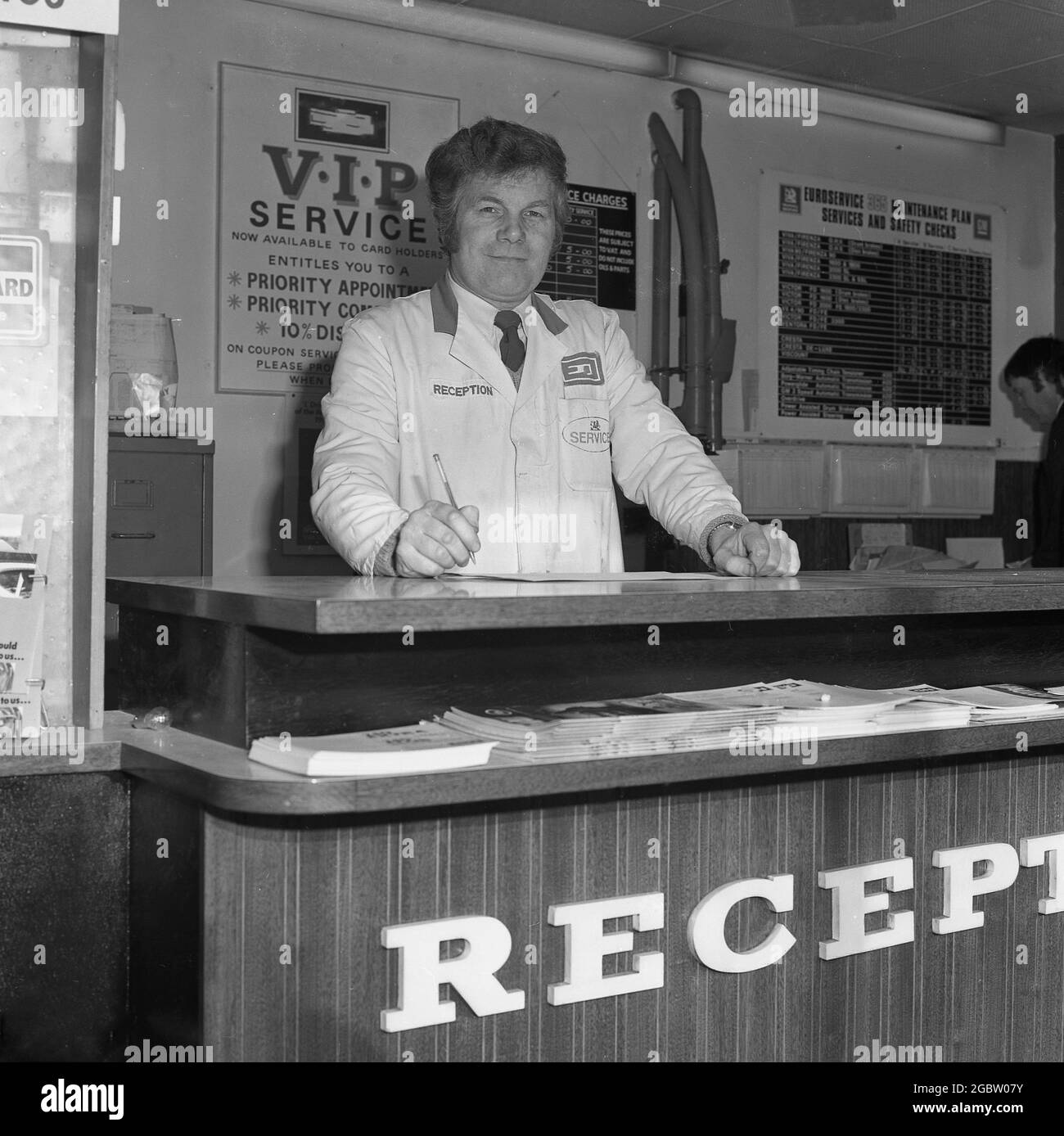 1975, historisch, ein leitender männlicher Mechaniker in einem weißen Mantel mit Firmenlogos, der am Schalter der Serviceannahme einer Garage in Croydon, England, Großbritannien, stand. Stockfoto