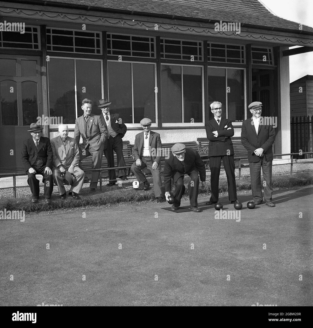 Historische Männer der 1960er Jahre in formellen Anzügen spielen Rasenbowling, während andere Männer vor dem Clubhaus im Bowlingclub in Kelty, Fife, Schottland, Großbritannien, zuschauen. In dieser Zeit würden Männer für fast alle ihre Aktivitäten, bei der Arbeit, am Strand und wie hier zu sehen, zum Sport und einem gemütlichen Bowellspiel, Anzüge tragen. Stockfoto