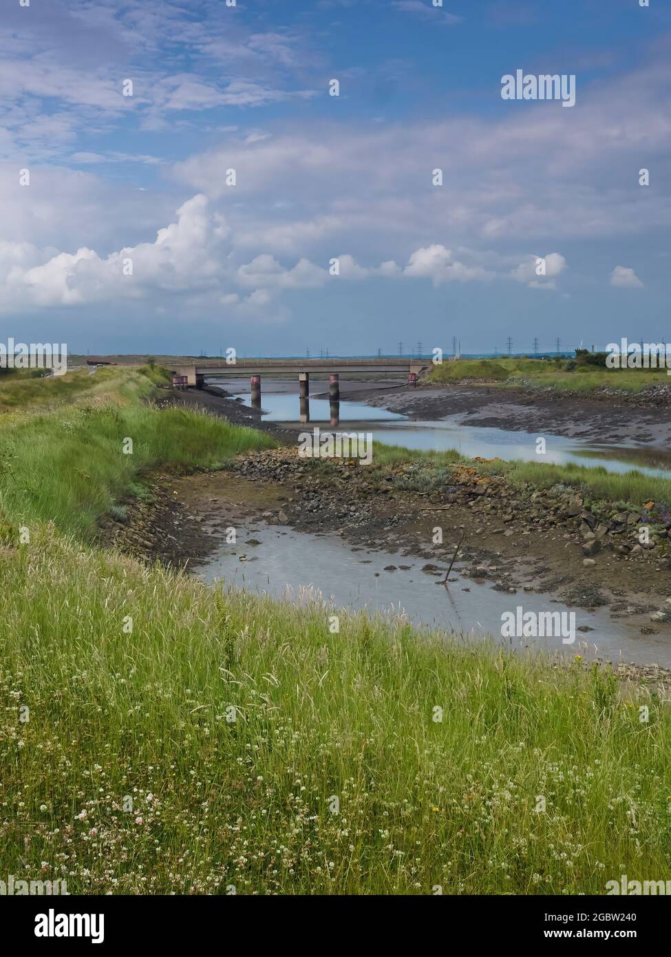 Ein Grasland und ein zerklüfteter Flussufer führen zu einer Brücke unter der riesigen und komplizierten Wolkenlandschaft voller Raum und Licht. Stockfoto