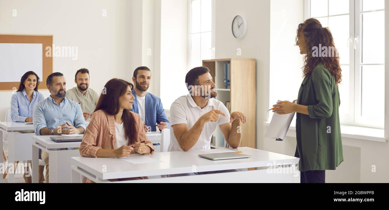 Coach, der mit einer Gruppe glücklicher Menschen diskutiert, die an Schreibtischen im Klassenzimmer sitzen Stockfoto