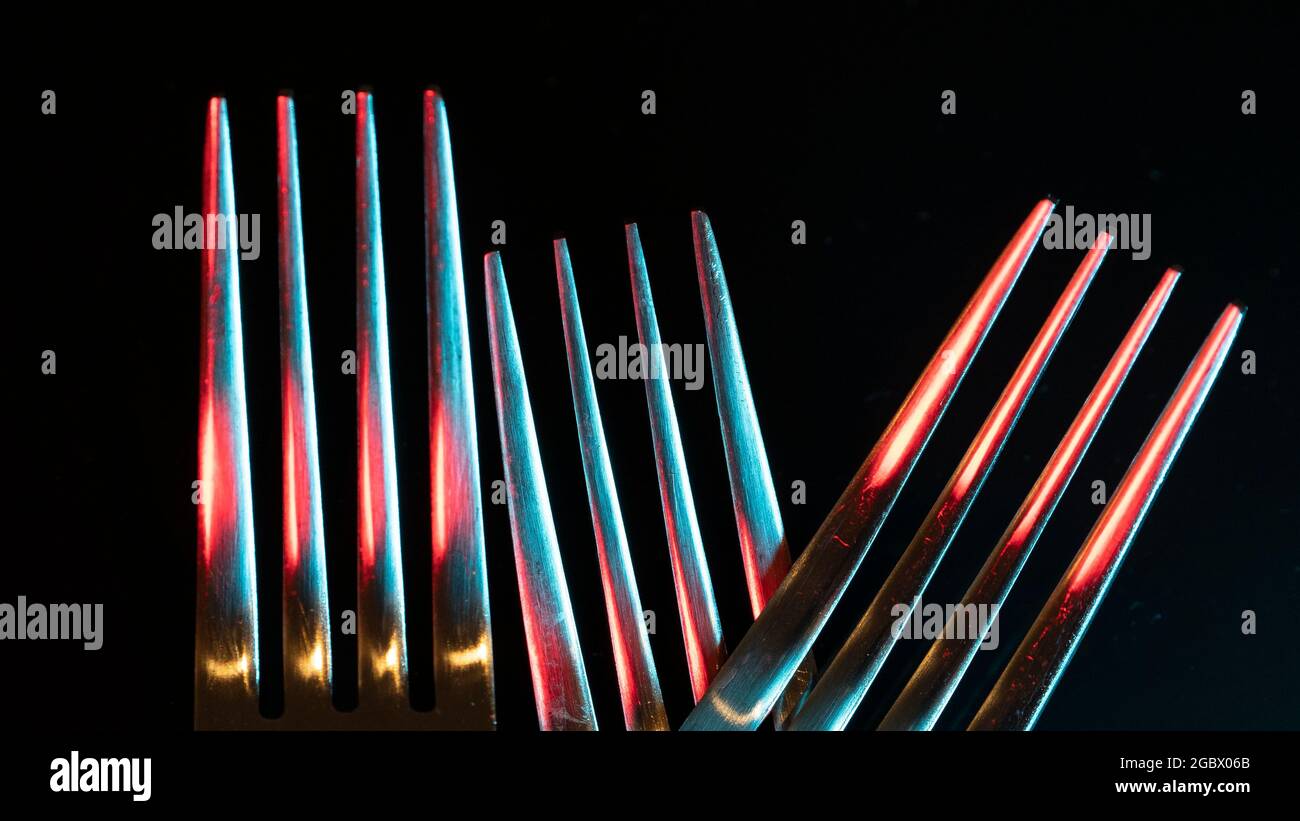 Nahaufnahme von drei glänzenden Gabeln auf schwarzem Hintergrund, die von farbigen Lichtern beleuchtet werden und ein abstraktes Bild bilden Stockfoto