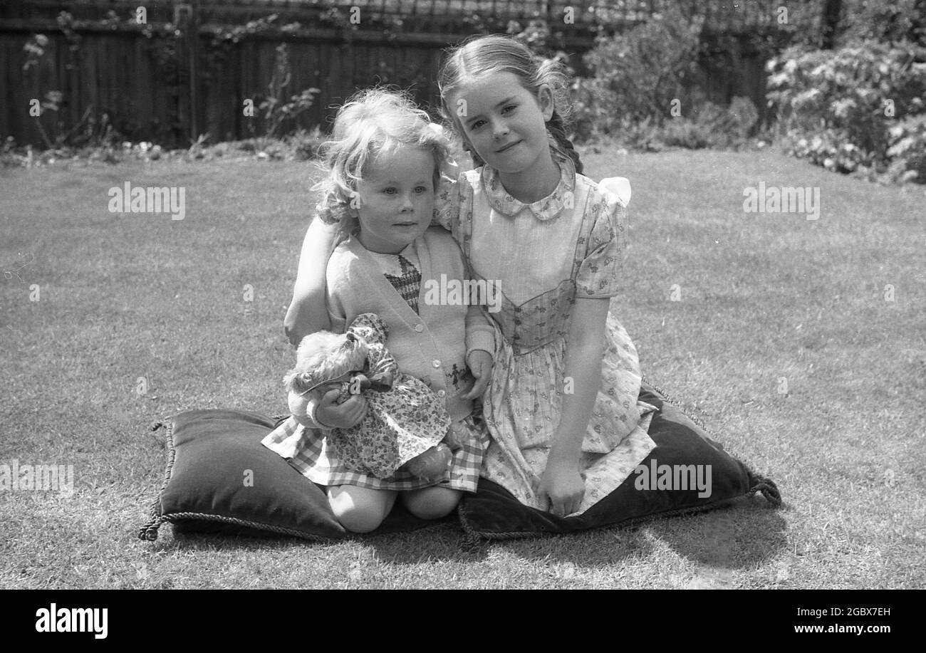 1960er Jahre, historisch, sommerlich und draußen auf einem Gartenrasen, ein junges Mädchen mit dem Arm um ihre Schwester, die beide auf Kissen knieten, um ihr Foto zu machen. Das jüngste Mädchen hält ihr weiches Spielzeug, einen kleinen Teddybären mit einem blumengemusterten Baumwolltop, England, Großbritannien. Stockfoto