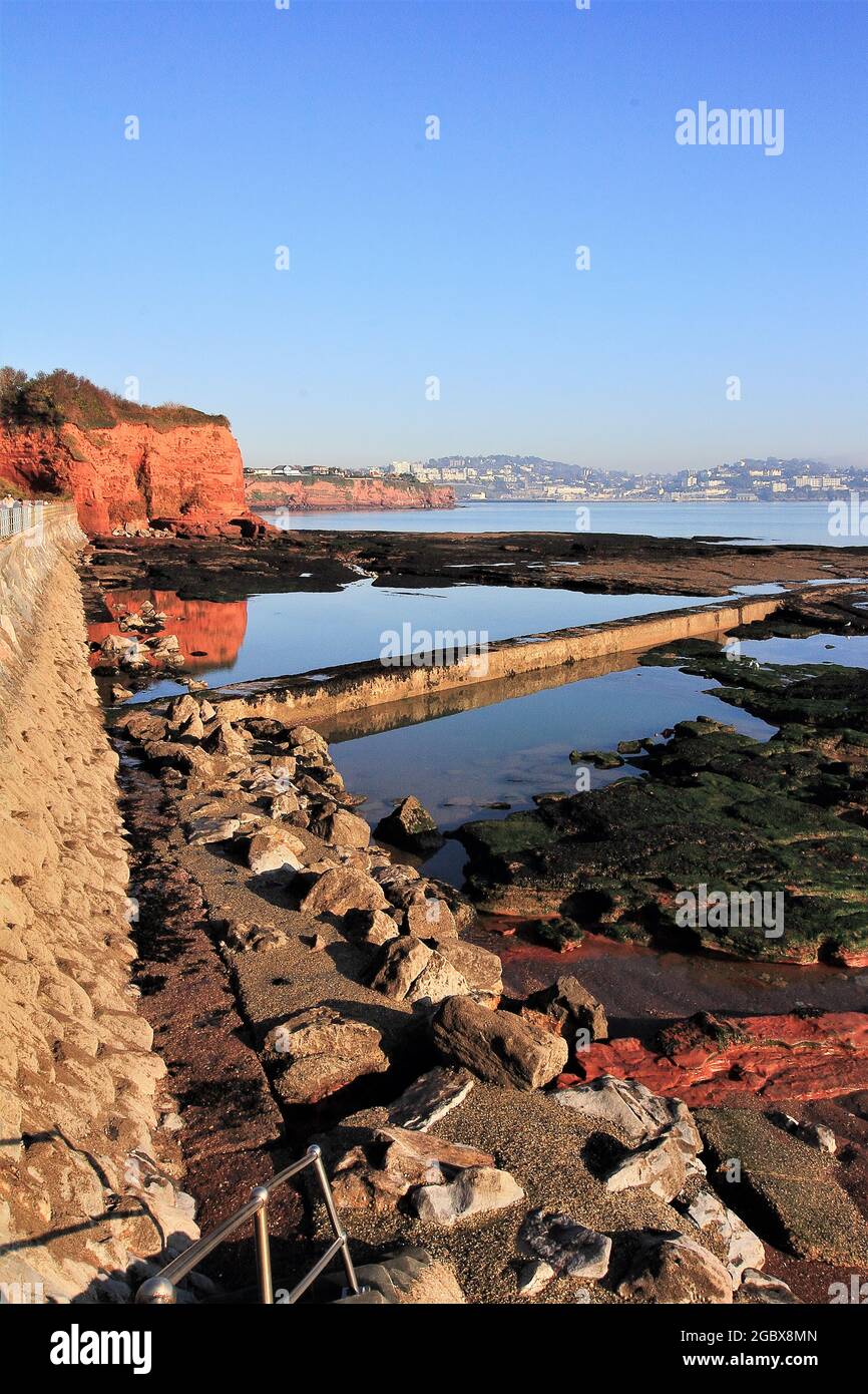 Ein Blick von Preston Sands, Paignton, Torbay, auf die roten Sandsteinklippen und die Reflexion im stillen Wasser eines Felsenpools, weiter um die Küste ist Stockfoto