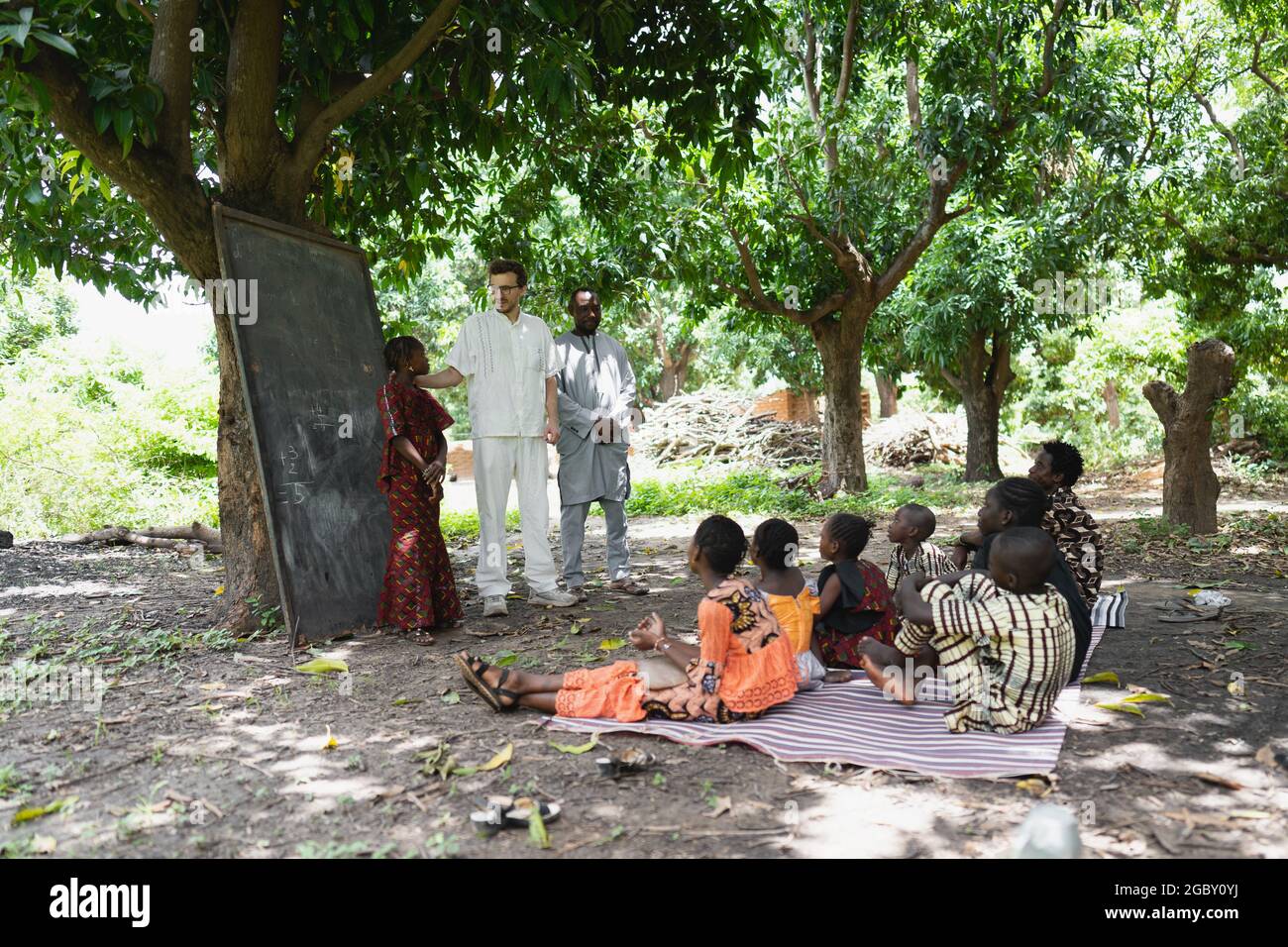 In diesem Bild unterrichtet ein großer weißer Freiwilliger eine Gruppe von Kindern in einem Sommerlager unter freiem Himmel in einem abgelegenen Dorf in Westafrika Stockfoto