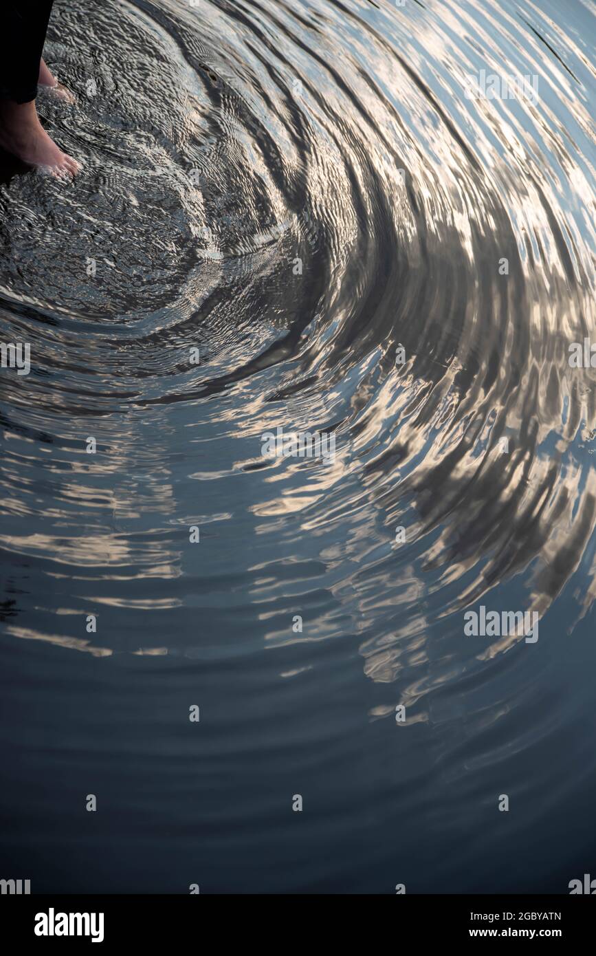 Die Füße einer Person im Wasser erzeugen Wellen an der Seenoberfläche mit reflektierten Himmel Stockfoto
