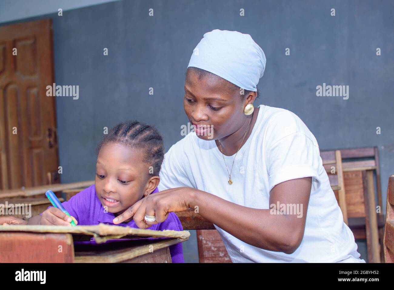 Afrikanische nigrische Mutter oder Lehrerin, die mit ihrem Kind in einem Klassenzimmer sitzt und ihr beim Studium hilft, in ihrer Schule hervorragende Leistungen zu erzielen Stockfoto