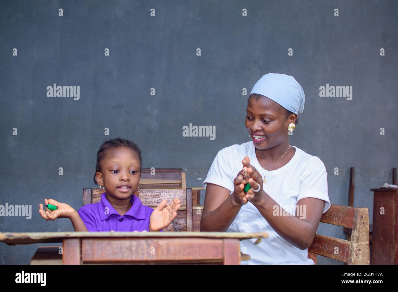 Afrikanische nigrische Mutter oder Lehrerin, die mit ihrem Kind in einem Klassenzimmer sitzt und ihr beim Studium hilft, in ihrer Schule hervorragende Leistungen zu erzielen Stockfoto